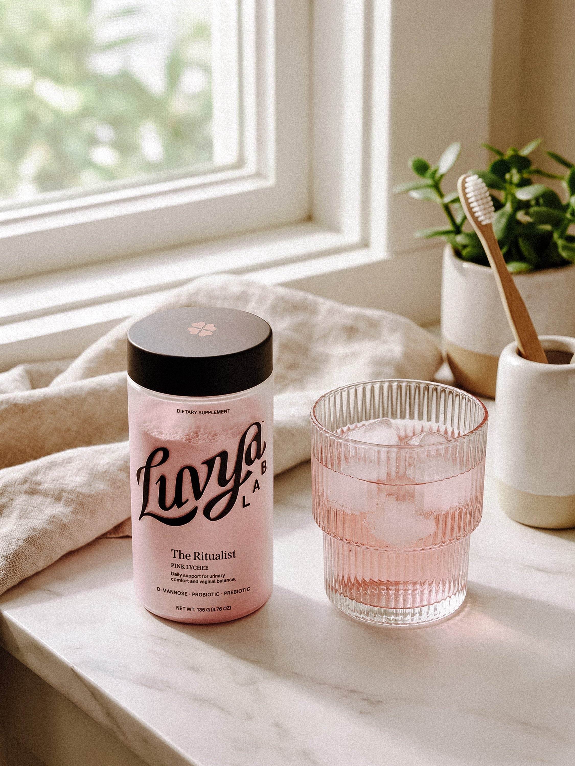A Luvya Lab jar on a sunlit windowsill beside a ribbed glass of the pink drink