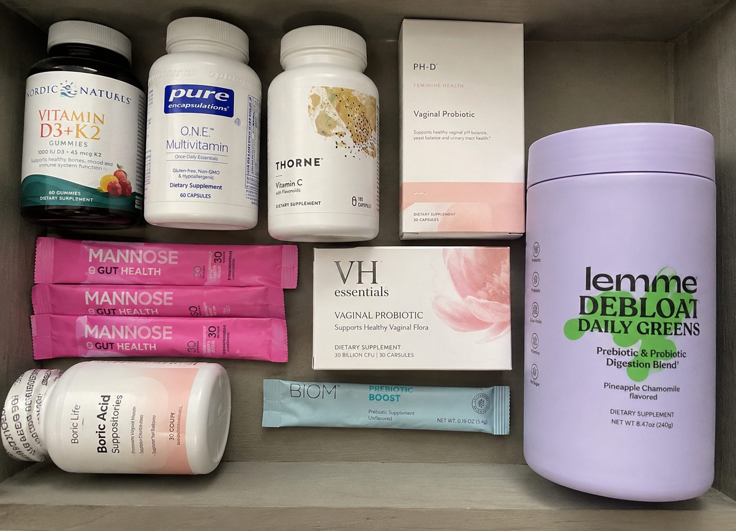 An open cardboard drawer holding four separate supplement products: a vitamin D3 plus K2 bottle, a multivitamin, a vitamin C bottle, a boxed vaginal probiotic, pink D-Mannose stick packs, a probiotic sachet box and a large lavender daily greens tub