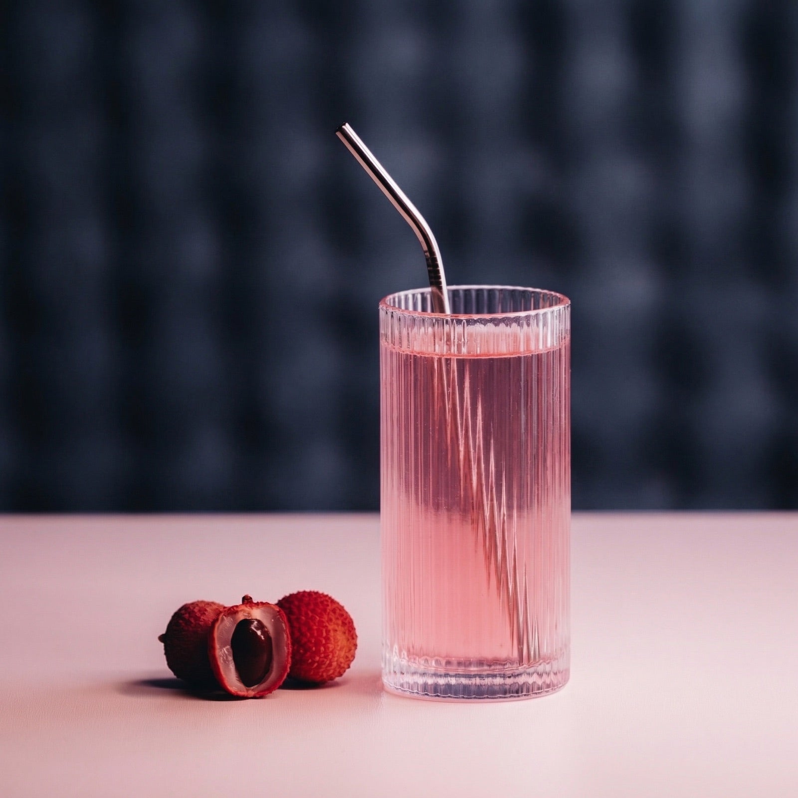 A ribbed glass of pink drink with a metal straw and lychee on a pink tabletop