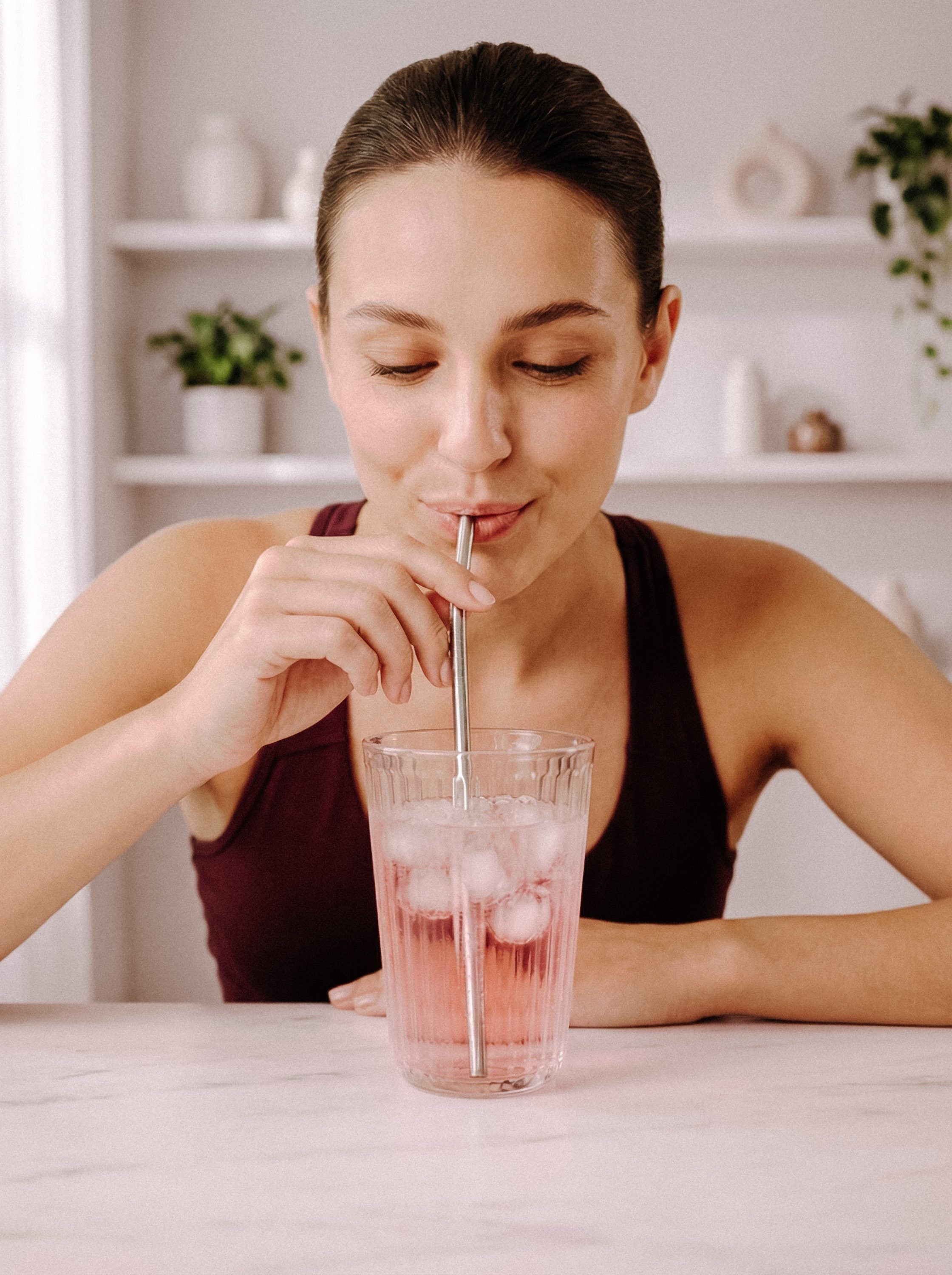 A woman leaning on a kitchen counter sipping a tall pink iced drink through a straw