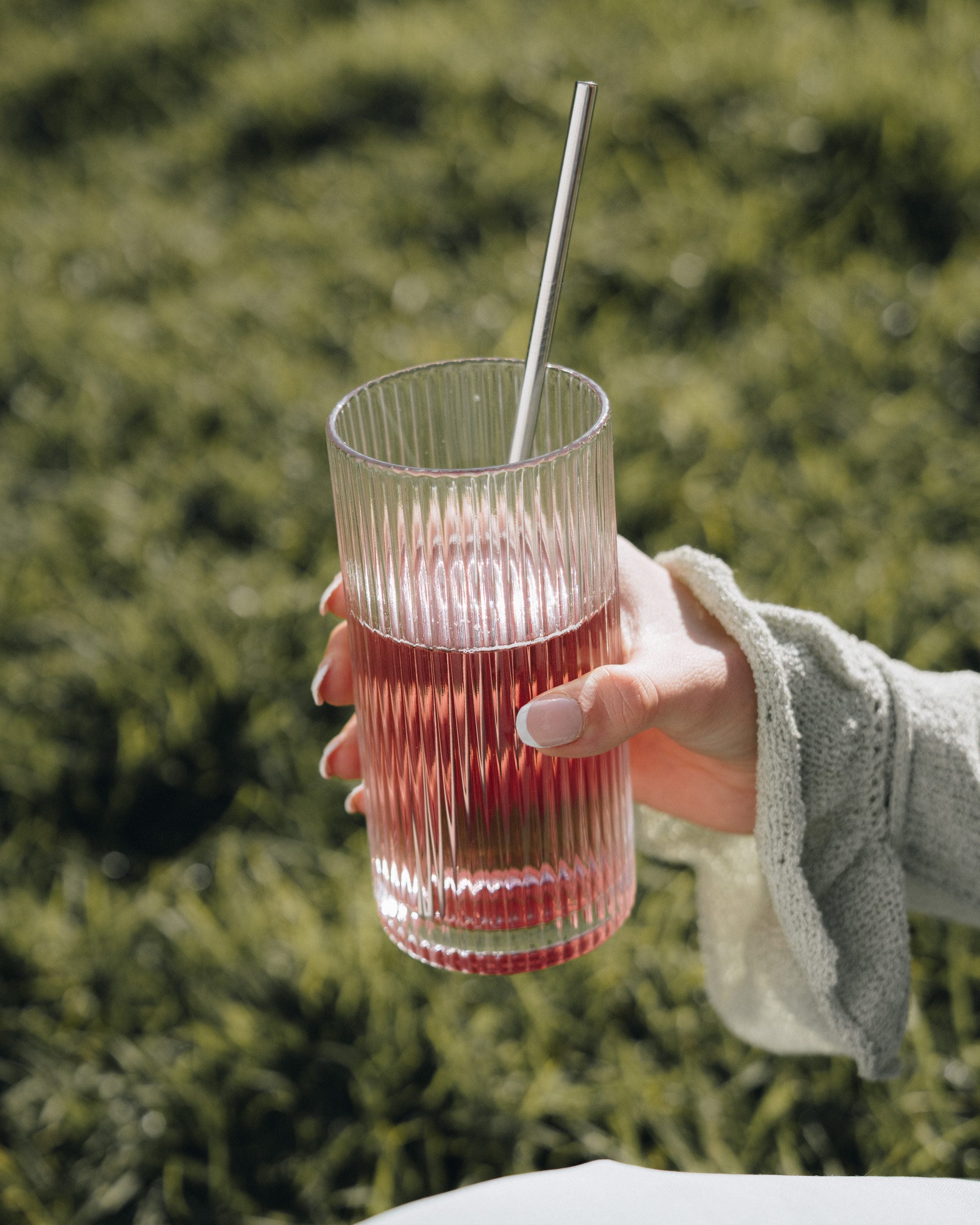 A hand holding a ribbed glass of pale pink Luvya Lab drink outdoors in front of green foliage